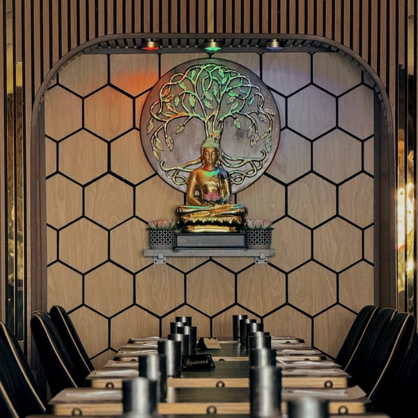 Restaurant interior with a golden Buddha statue, tree of life art, and long tables with chairs.
