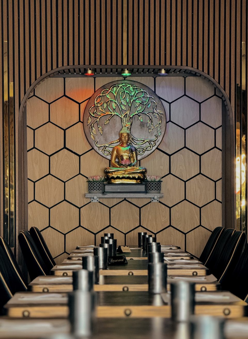 Restaurant interior with a golden Buddha statue, tree of life art, and long tables with chairs.