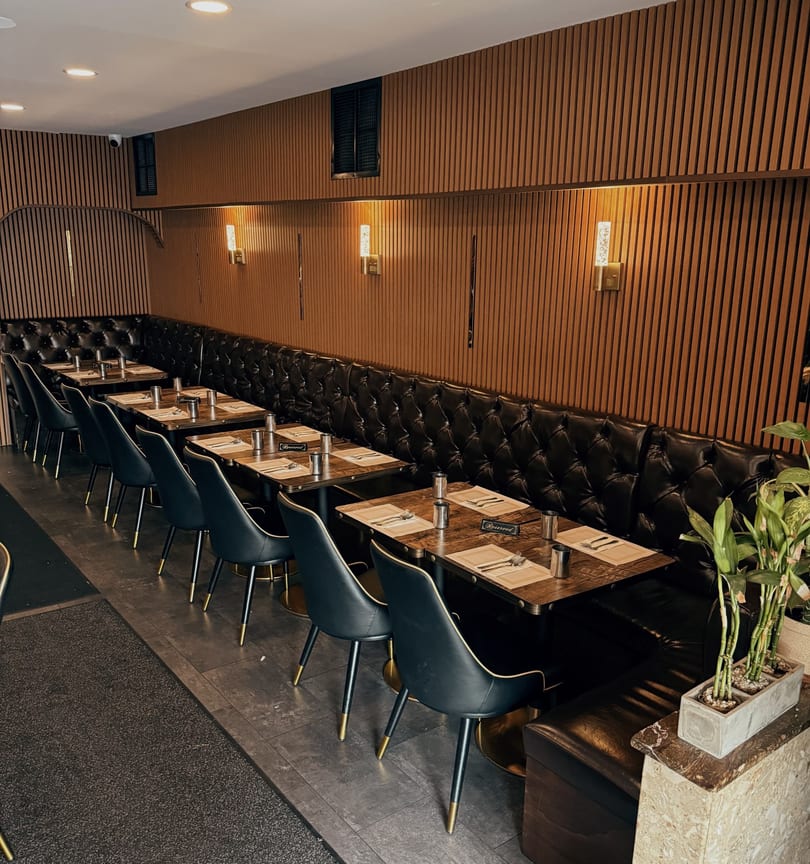 A long row of set tables with black chairs and a tufted leather banquette against a wood-paneled wall.