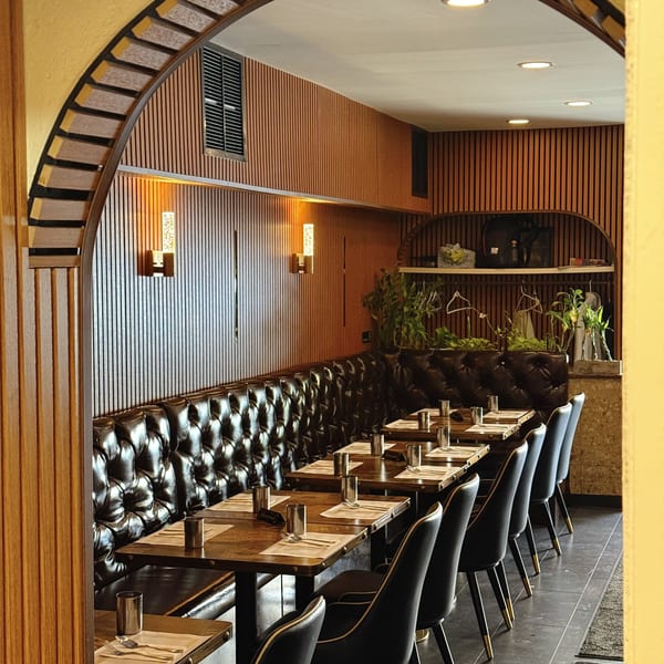 A restaurant dining room with dark tufted leather banquettes, wooden tables, and black chairs with gold accents.