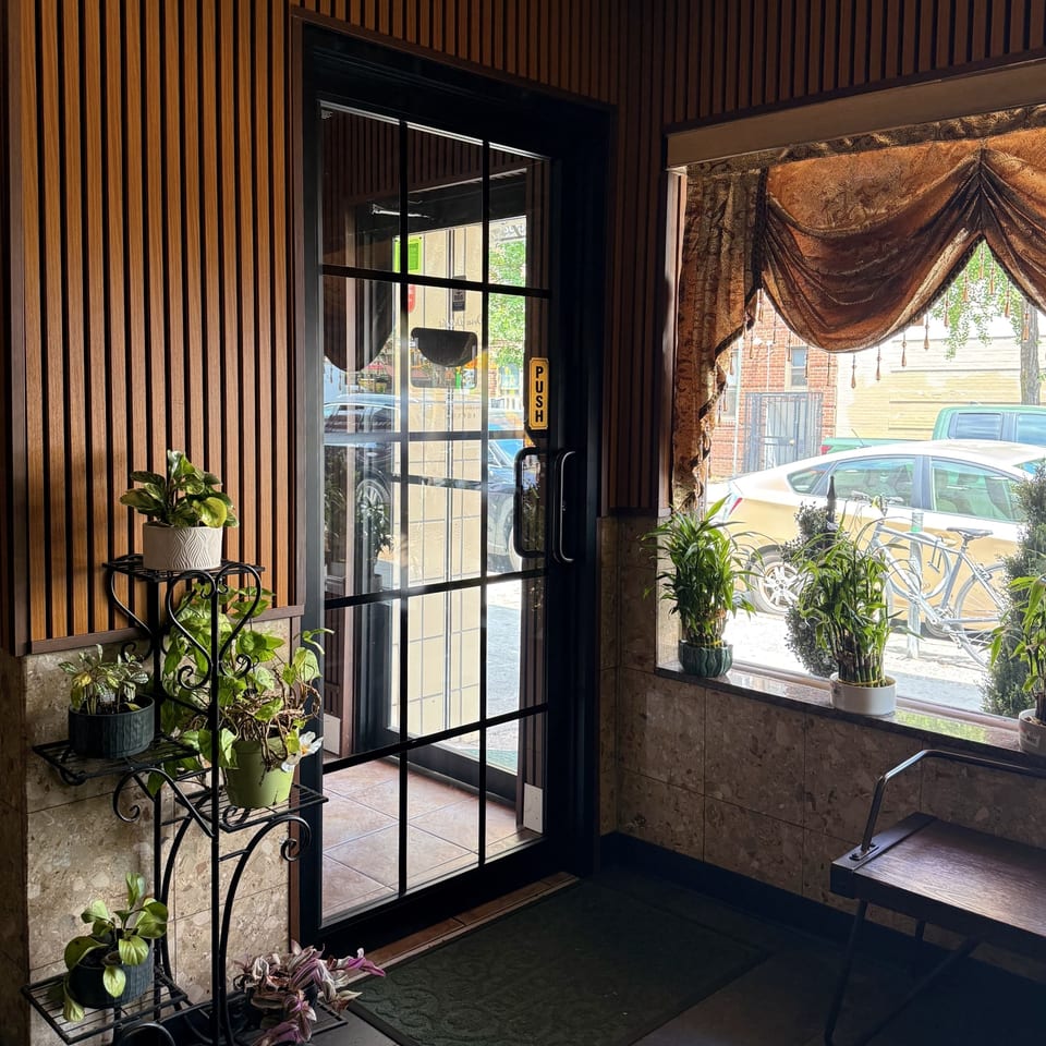 Restaurant entrance with wood paneling, glass door, potted plants, and a window with ornate curtains.