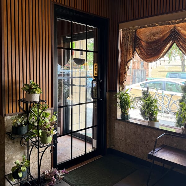 Restaurant entrance with wood-paneled walls, potted plants, a glass door, and a window with ornate curtains.