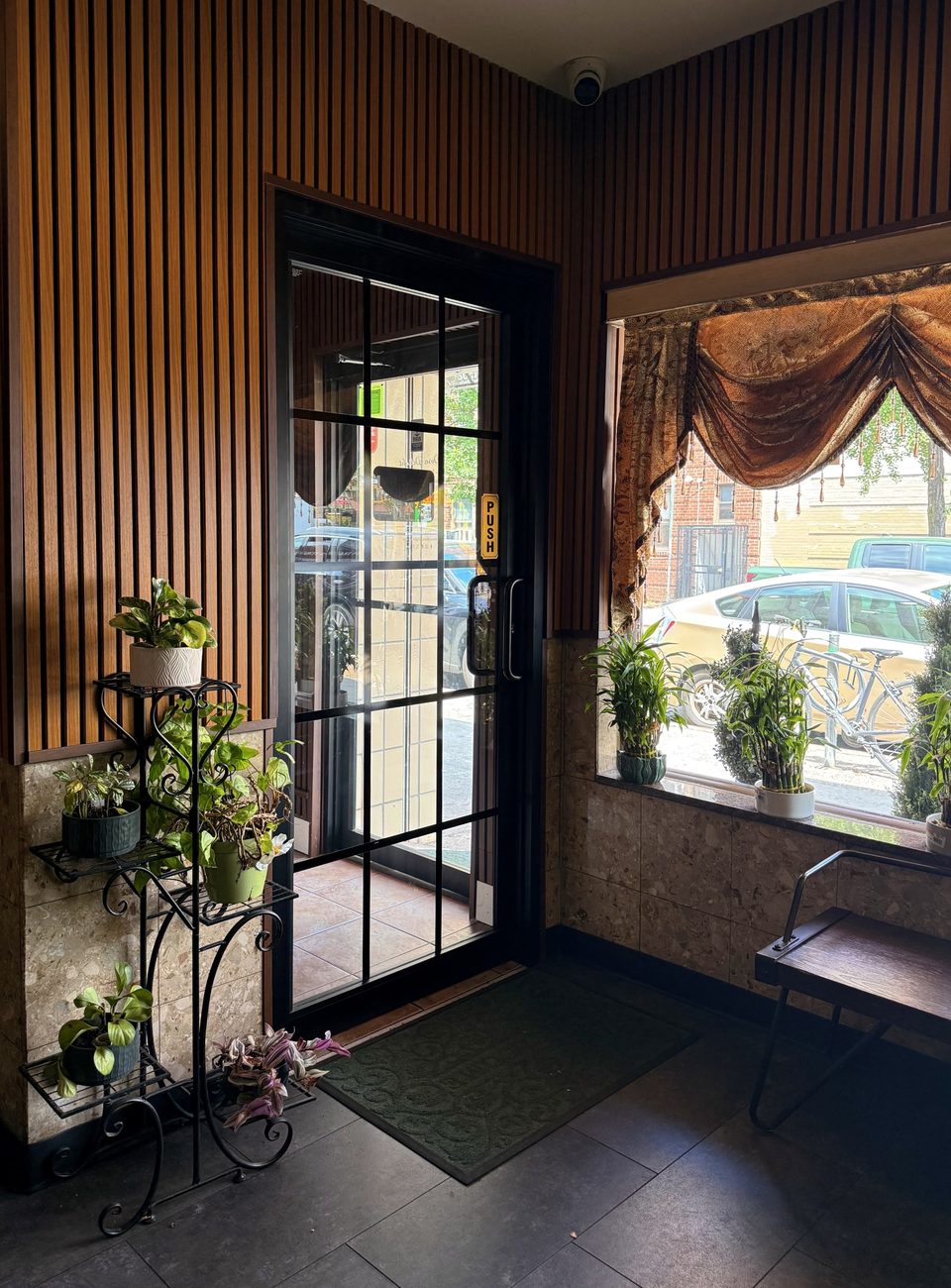 Restaurant entrance with wood-paneled walls, potted plants, a glass door, and a window with ornate curtains.